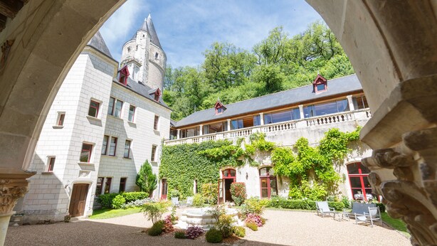 Un patio con un arco de piedra, un edificio con una torre y un jardín con plantas y una fuente.