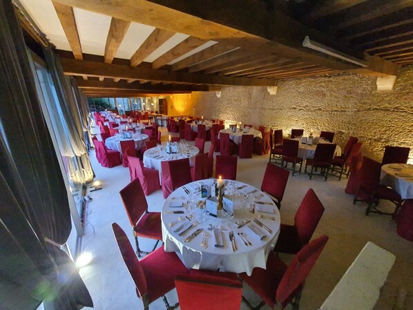 A spacious dining area with round tables and red chairs, set against a stone wall and wooden beams.