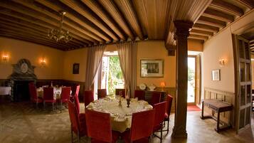A dining area with a round table set for six, surrounded by red upholstered chairs. A fireplace is visible in the background, and the room has wooden beams on the ceiling.