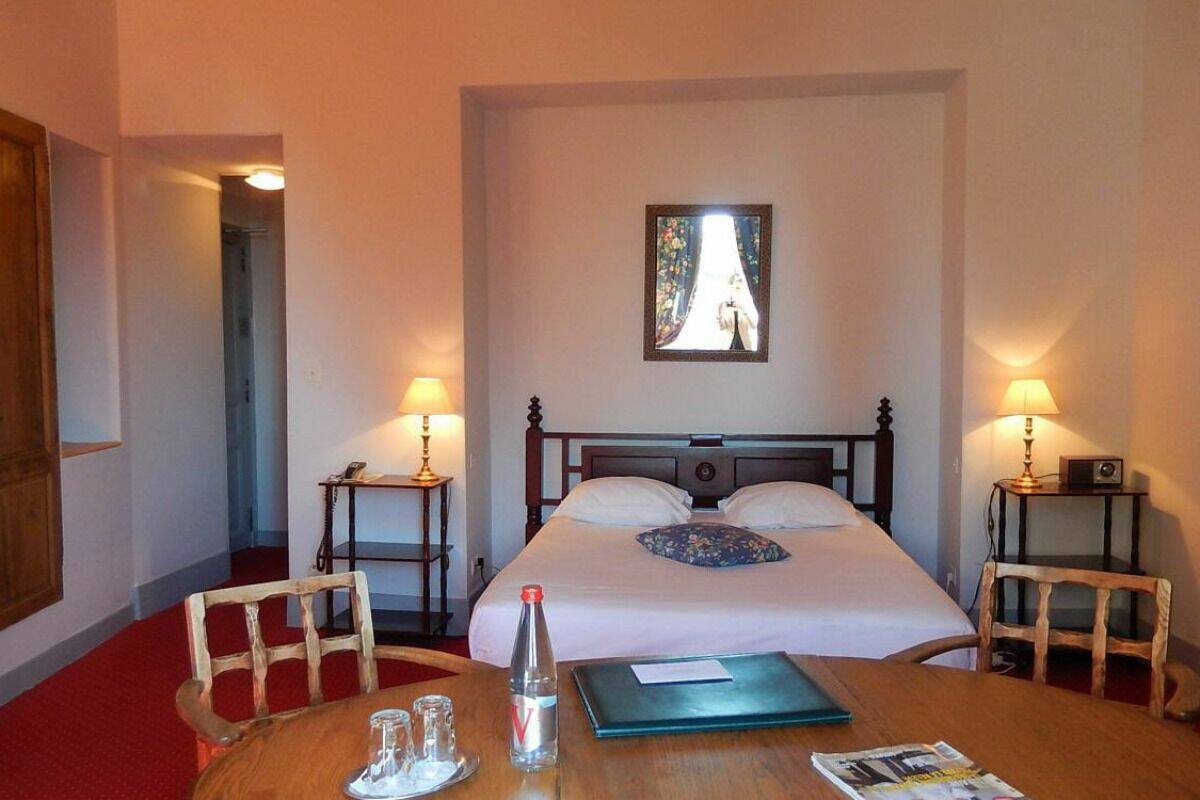 Une chambre d’hôtel avec un lit, deux tables de chevet avec des lampes, une chaise en bois, une table avec une bouteille, des verres et un livre.