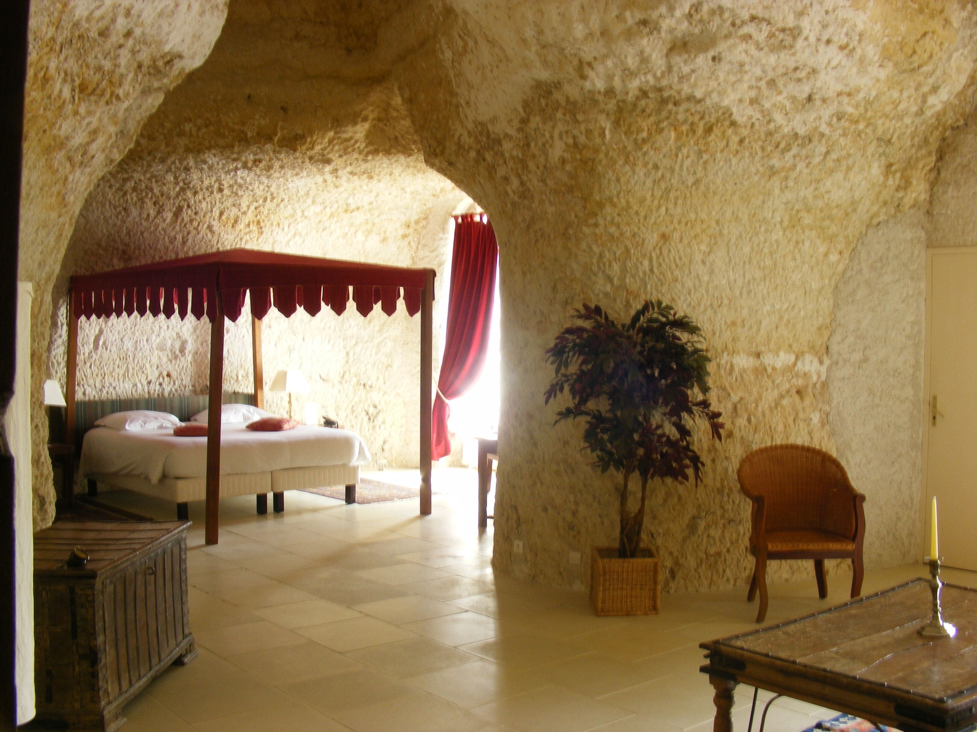 A bedroom with a canopy bed, a potted plant, a chair, and a table with a candle.