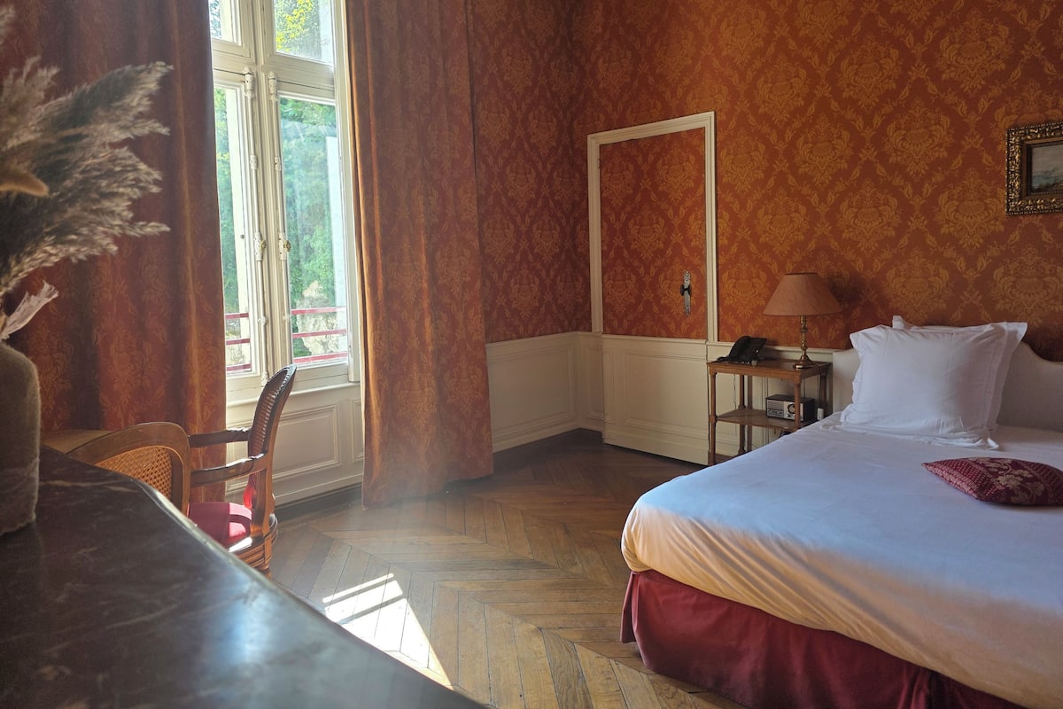 Une chambre d’hôtel avec un lit, un bureau, une chaise, une lampe et une fenêtre avec des rideaux.
