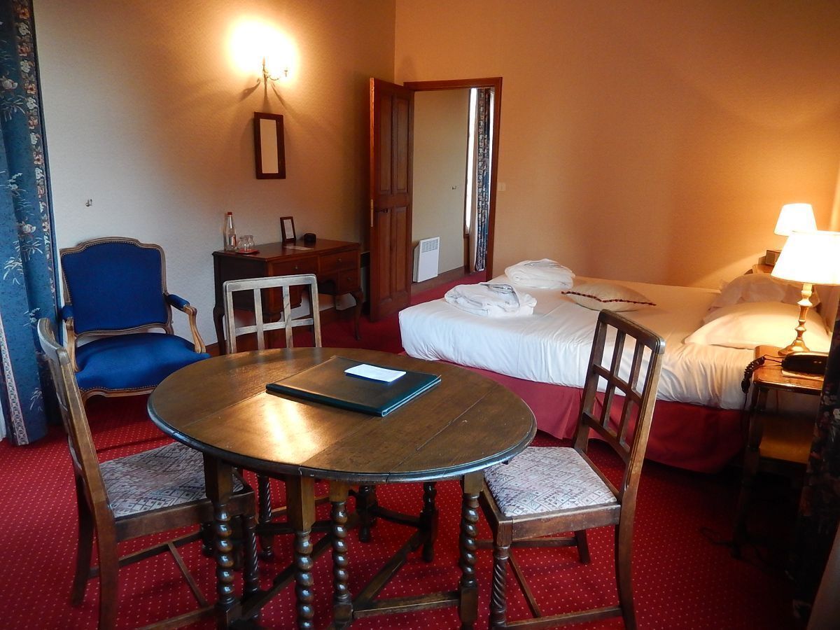 Una habitación de hotel con una cama, una mesa redonda de madera con sillas, un escritorio, una silla, una lámpara y un espejo.