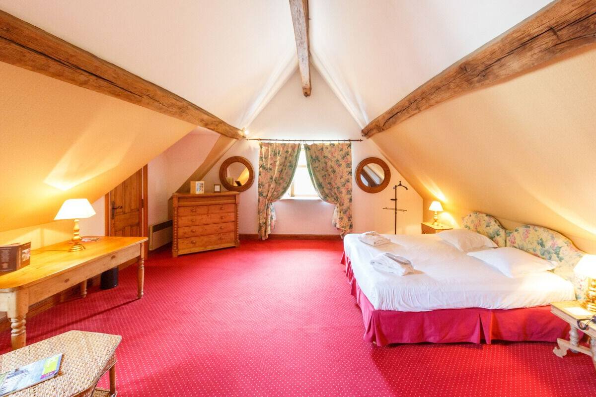 Une chambre confortable située sous les toits, avec un tapis rouge, des meubles en bois, un lit recouvert de draps blancs et une commode en bois.