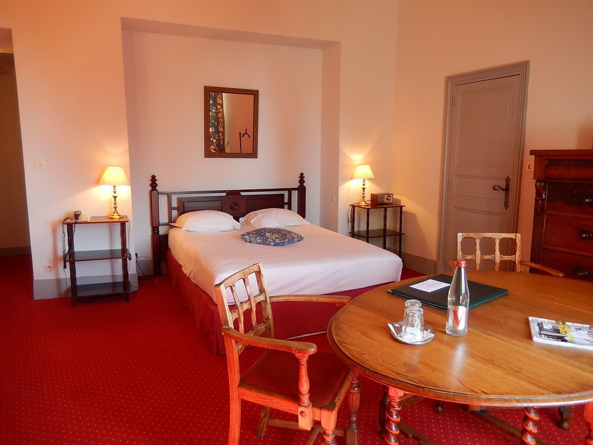 A hotel room with a double bed, two bedside tables with lamps, a wooden chair, a round wooden table with a bottle, a magazine, and a door.