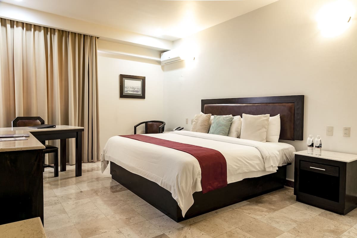 A hotel room with a bed, bedside table, desk, chair, and an air conditioning unit.