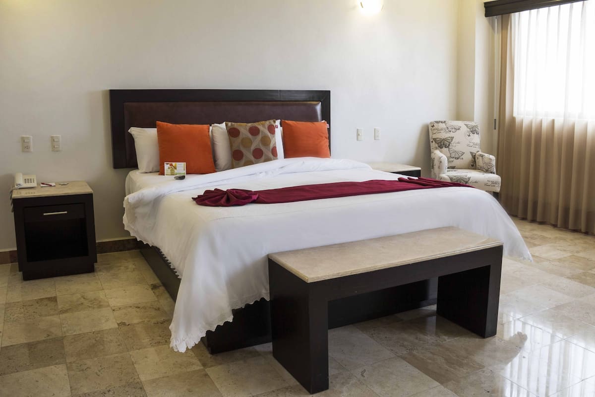 A neatly made bed with a headboard, a bedside table with a phone, a chair with a butterfly pattern, and a window with curtains.
