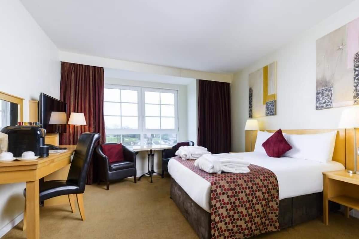 Una habitación de hotel con cama, escritorio, silla y ventana con cortinas.