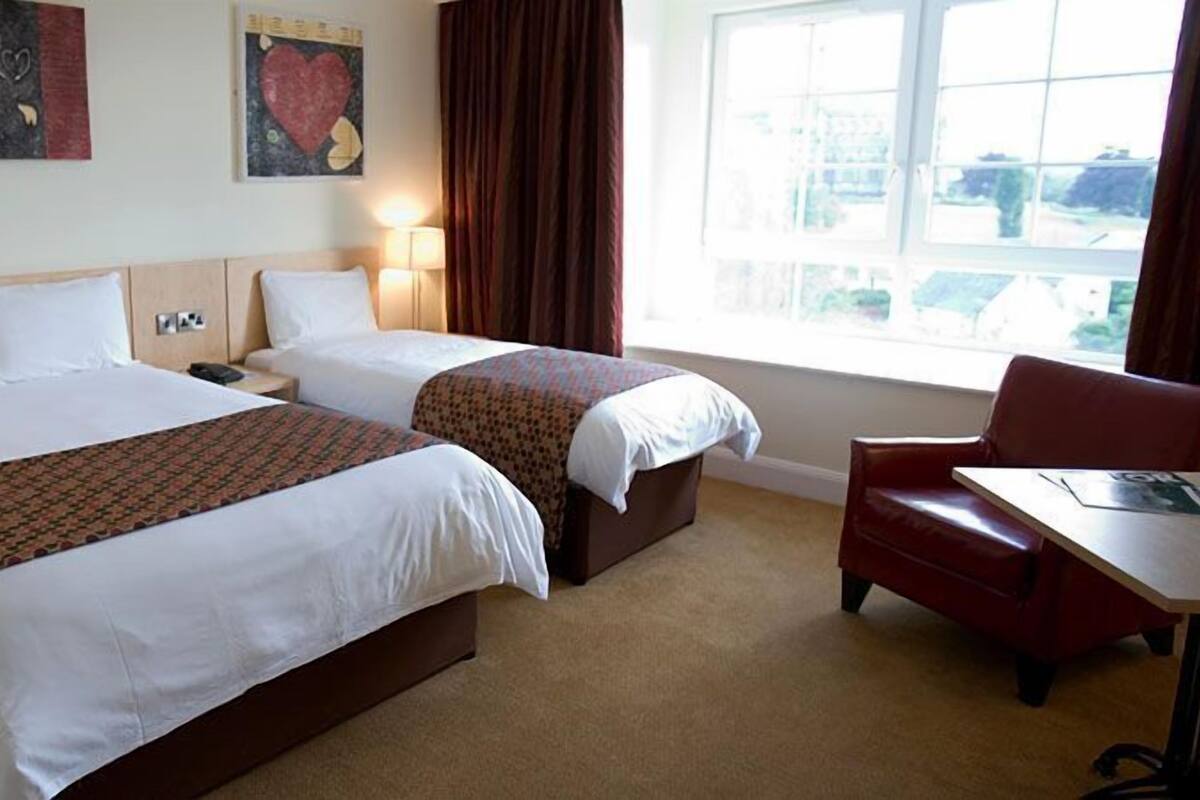 Habitación de hotel con dos camas, un escritorio y una silla. Hay dos mesitas de noche con lámparas. La habitación tiene un ventanal grande con cortinas.