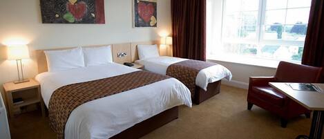A hotel room with two beds, a desk, and a chair. There are two bedside tables with lamps. The room has a large window with curtains.