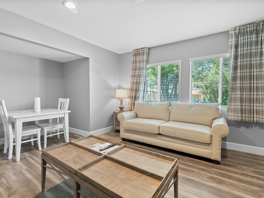 A living room with a beige sofa, a white dining table with chairs, a coffee table, and a lamp.