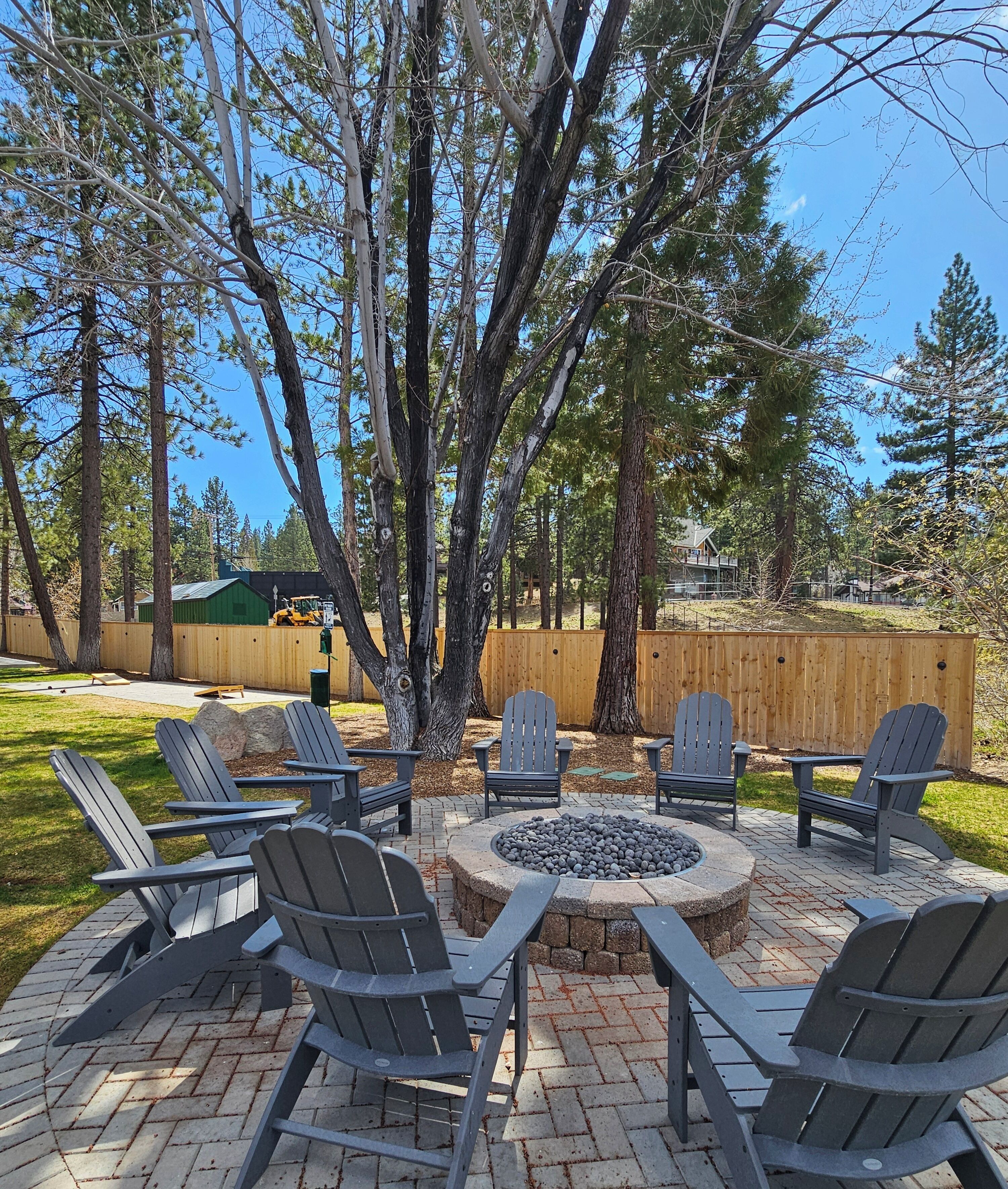 Une terrasse extérieure avec des fauteuils Adirondack et un foyer en pierre, entourée d’arbres et d’une clôture en bois.