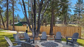 An outdoor patio with Adirondack chairs and a stone fire pit, surrounded by trees and a wooden fence.