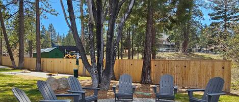 Une terrasse extérieure avec des fauteuils Adirondack et un foyer en pierre, entourée d’arbres et d’une clôture en bois.