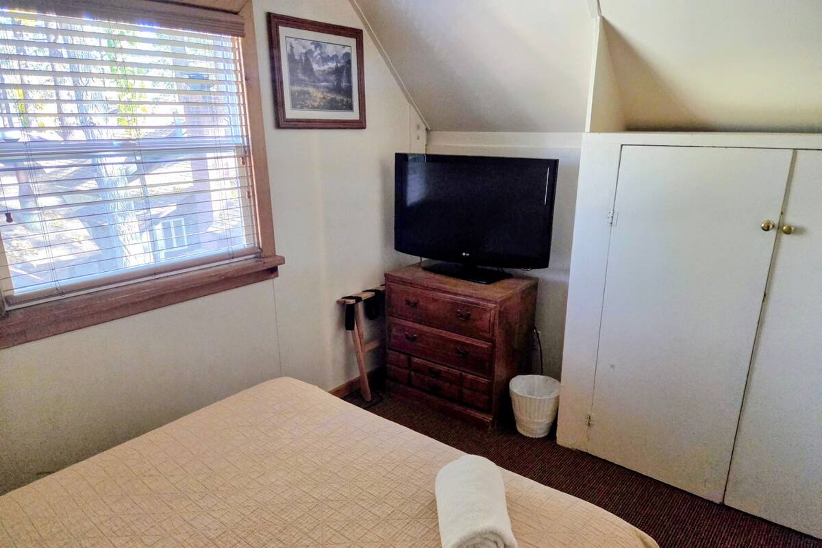 A bedroom with a bed, a television on a wooden dresser, a white wardrobe, and a window with blinds.