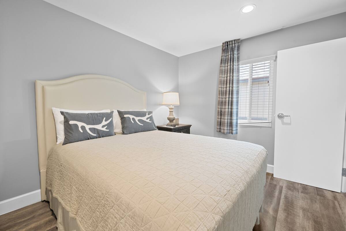 A bedroom with a large bed, a nightstand with a lamp, a window with blinds, and a door.