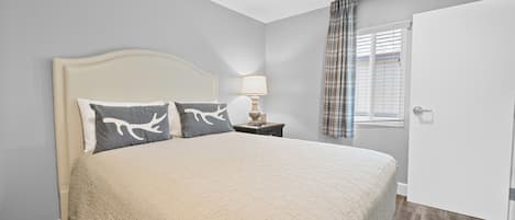 A bedroom with a large bed, a nightstand with a lamp, a window with blinds, and a door.
