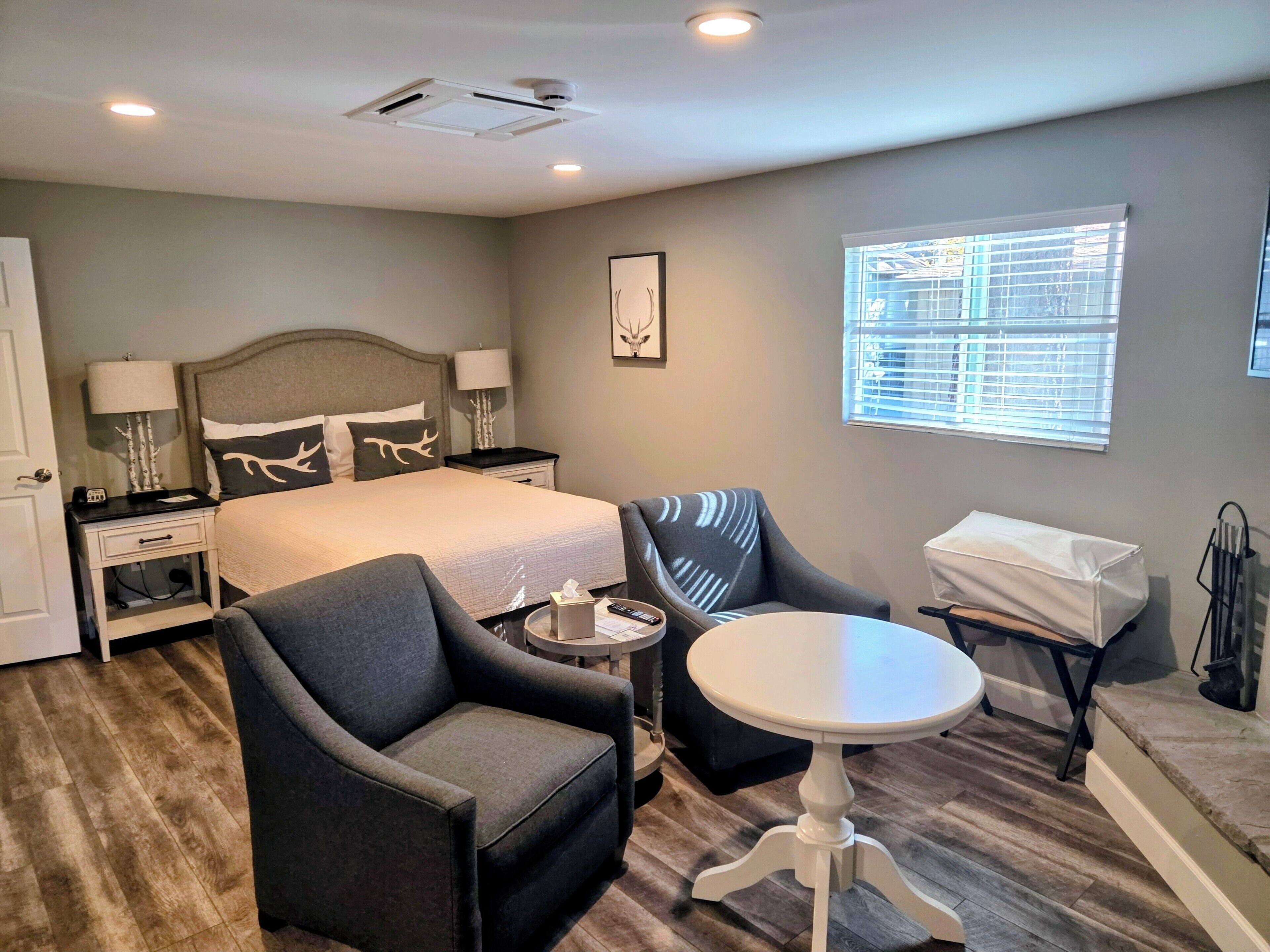 A hotel room with a bed, two armchairs, a small table, a bedside table, a lamp, a picture frame, and a window with blinds.