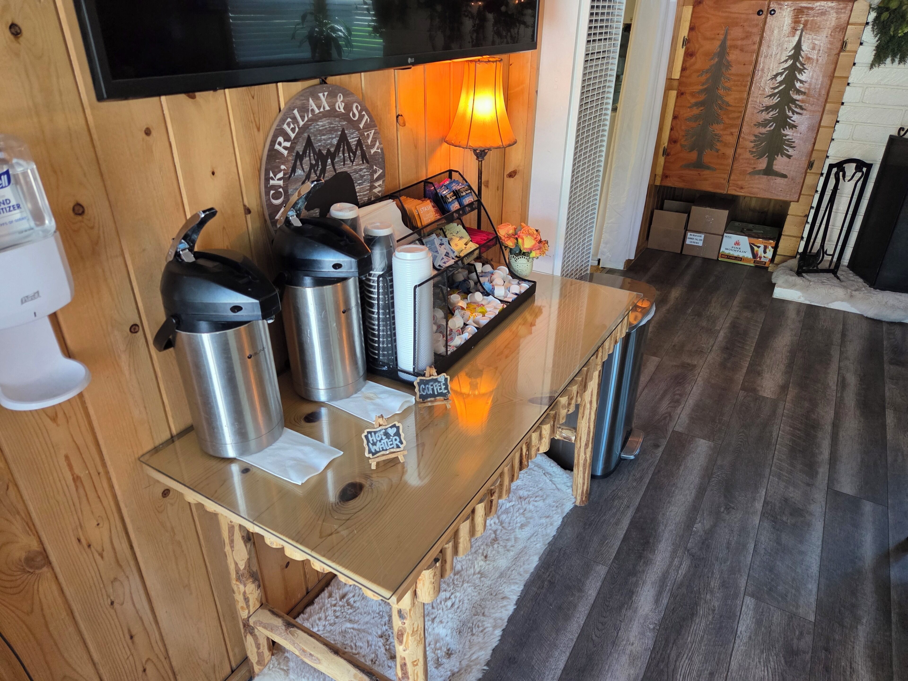 A wooden table with stainless steel thermos cups, a coffee station, and a wall-mounted TV.