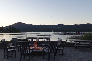 Un hogar de fuego rodeado de sillas y mesas con vista a un lago con barcos y montañas al fondo.