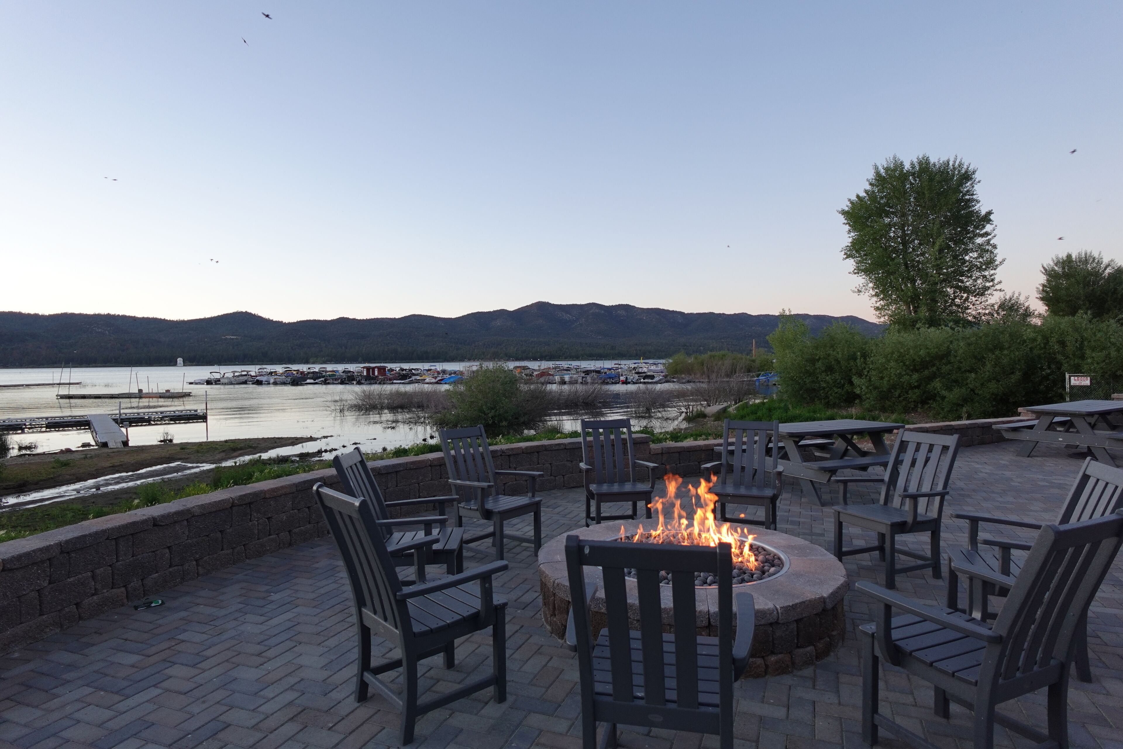 An outdoor seating area with a fire pit, chairs, and a view of a lake with boats and mountains in the background.