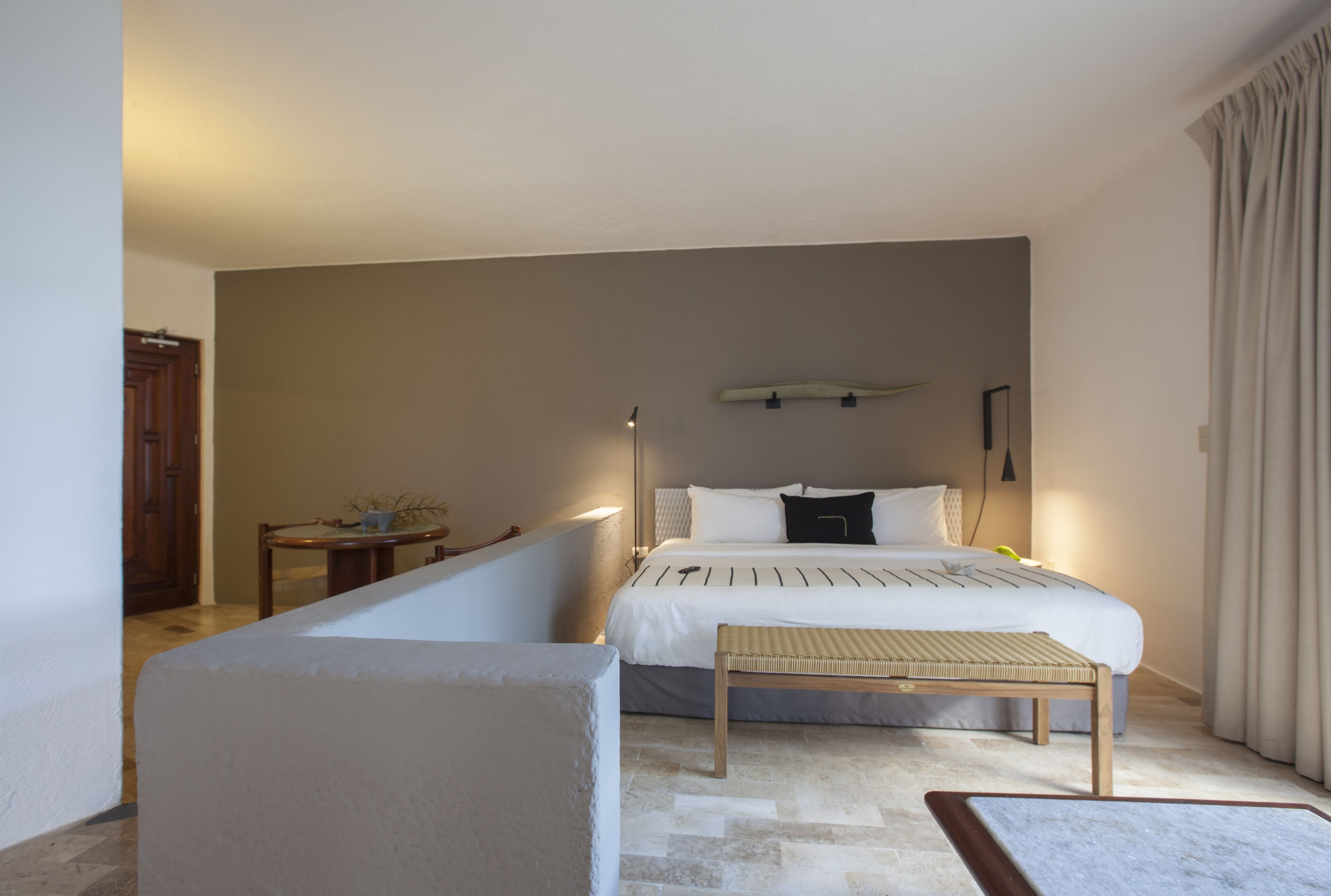 A hotel room with a bed, bedside table, bench, and a wall-mounted lamp.