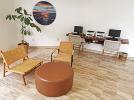 Una habitación con una planta grande en maceta, dos sillas de mimbre, un reposapiés marrón y un escritorio con impresoras y monitores.