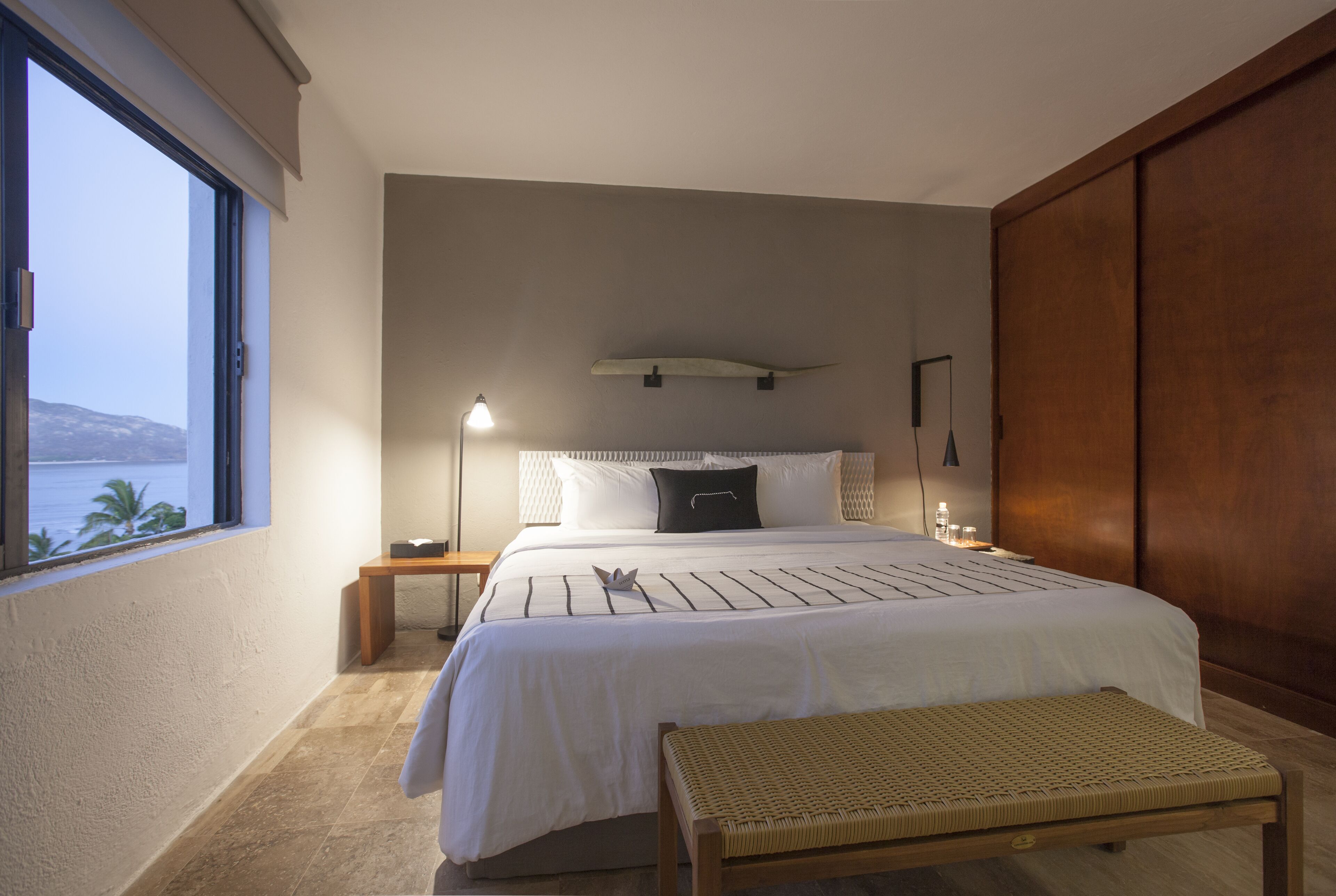 A bedroom with a large bed, a bedside table with a lamp, a bench, and a view of the sea.
