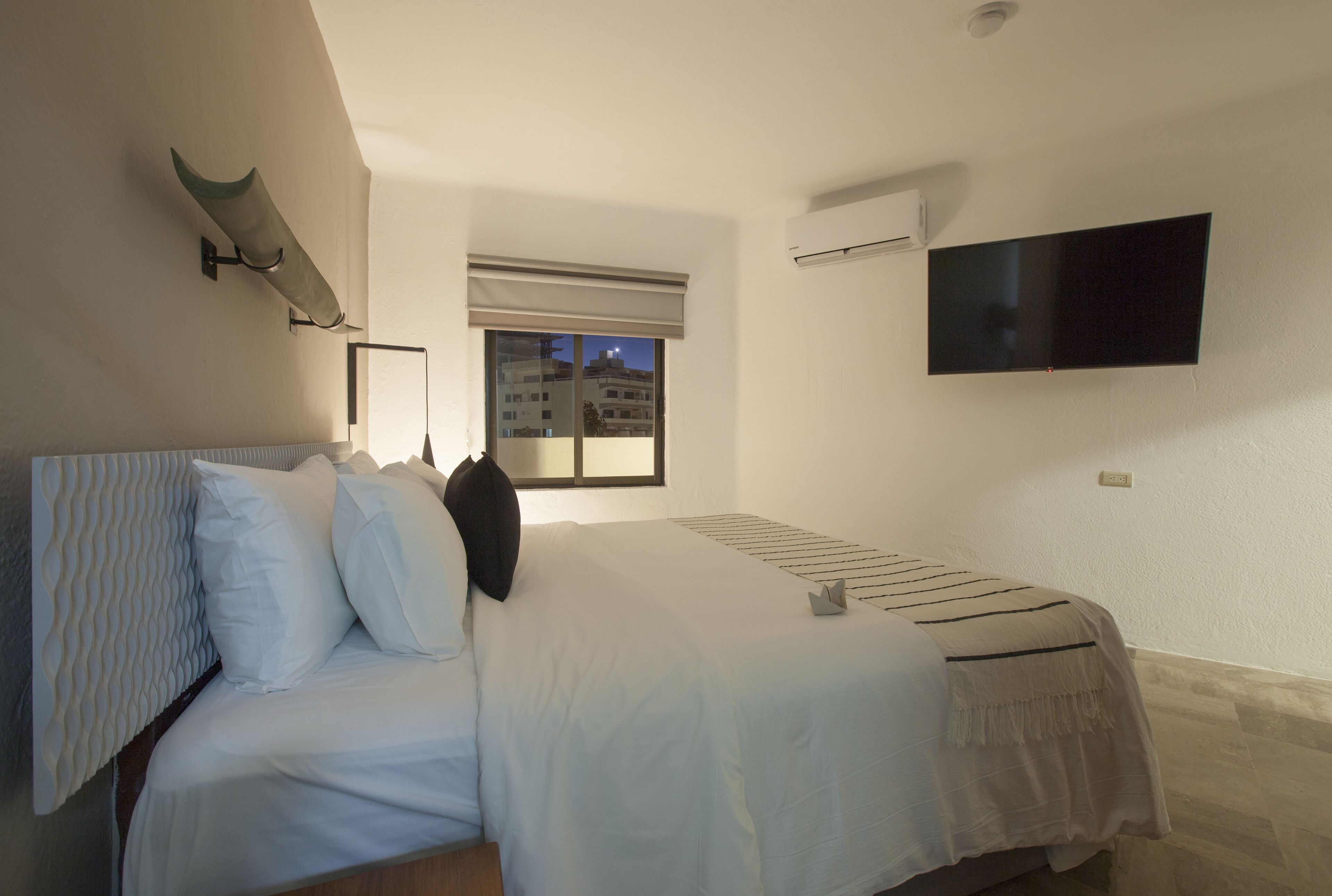 Un dormitorio con cama, televisor montado en la pared y ventana con vista a edificios.
