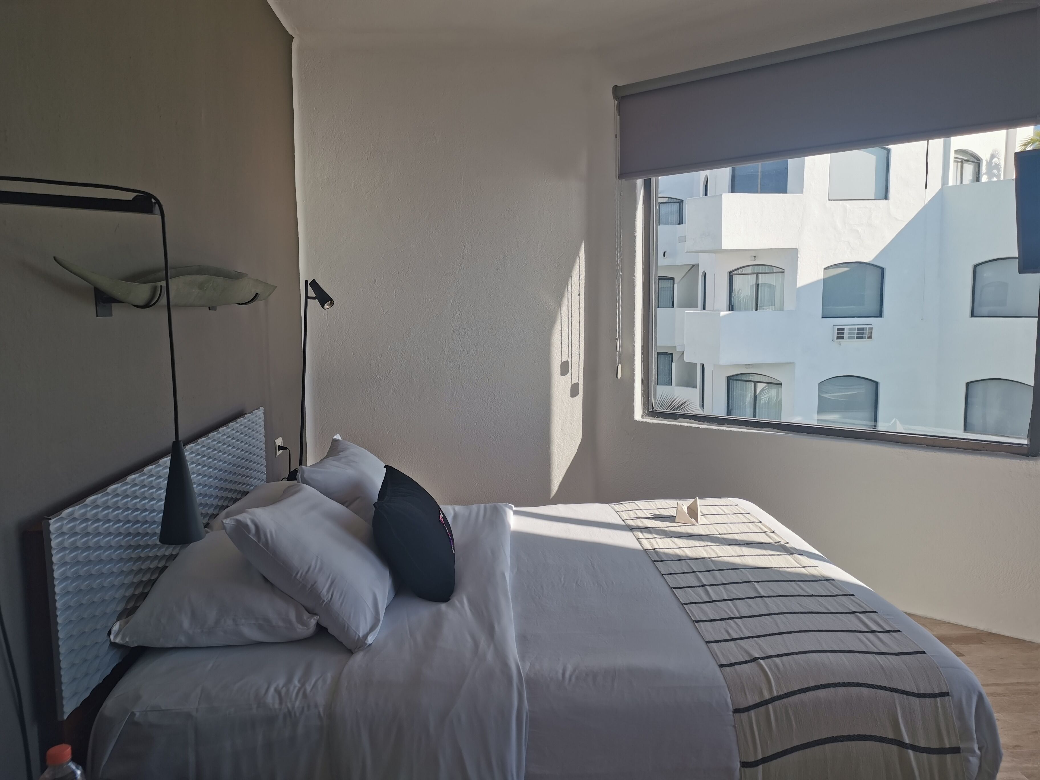 A modern hotel room with a large bed, a bedside lamp, and a view of a building through a window.