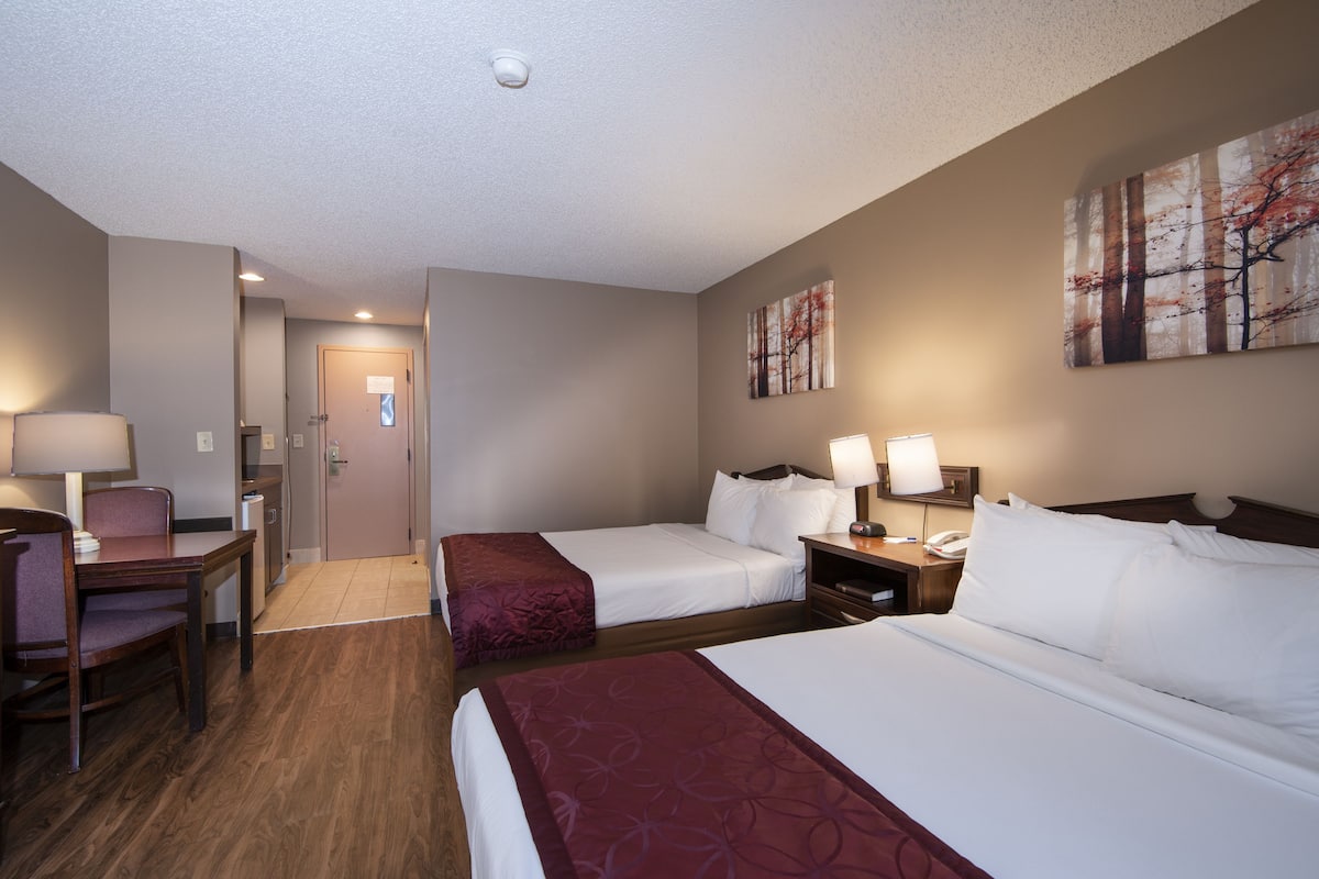 A hotel room with two beds, a desk, and a chair. There is a door leading to the bathroom and a hallway.