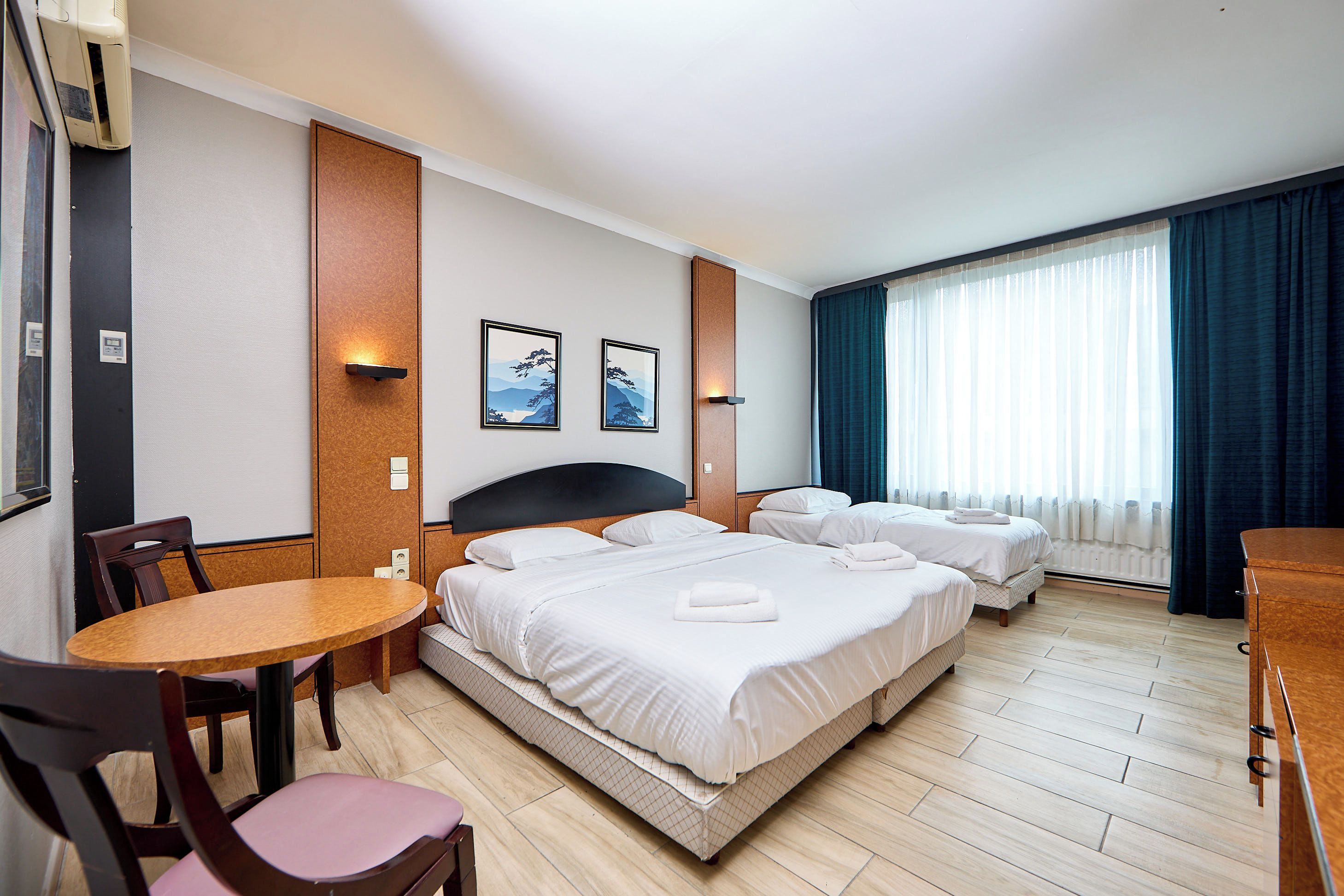 Une chambre d’hôtel avec un lit double, un lit simple, une table à manger et des chaises. Il y a deux tableaux encadrés au mur et une fenêtre avec des rideaux.