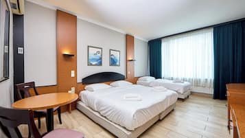 Une chambre d’hôtel avec un lit double, un lit simple, une table à manger et des chaises. Il y a deux tableaux encadrés au mur et une fenêtre avec des rideaux.