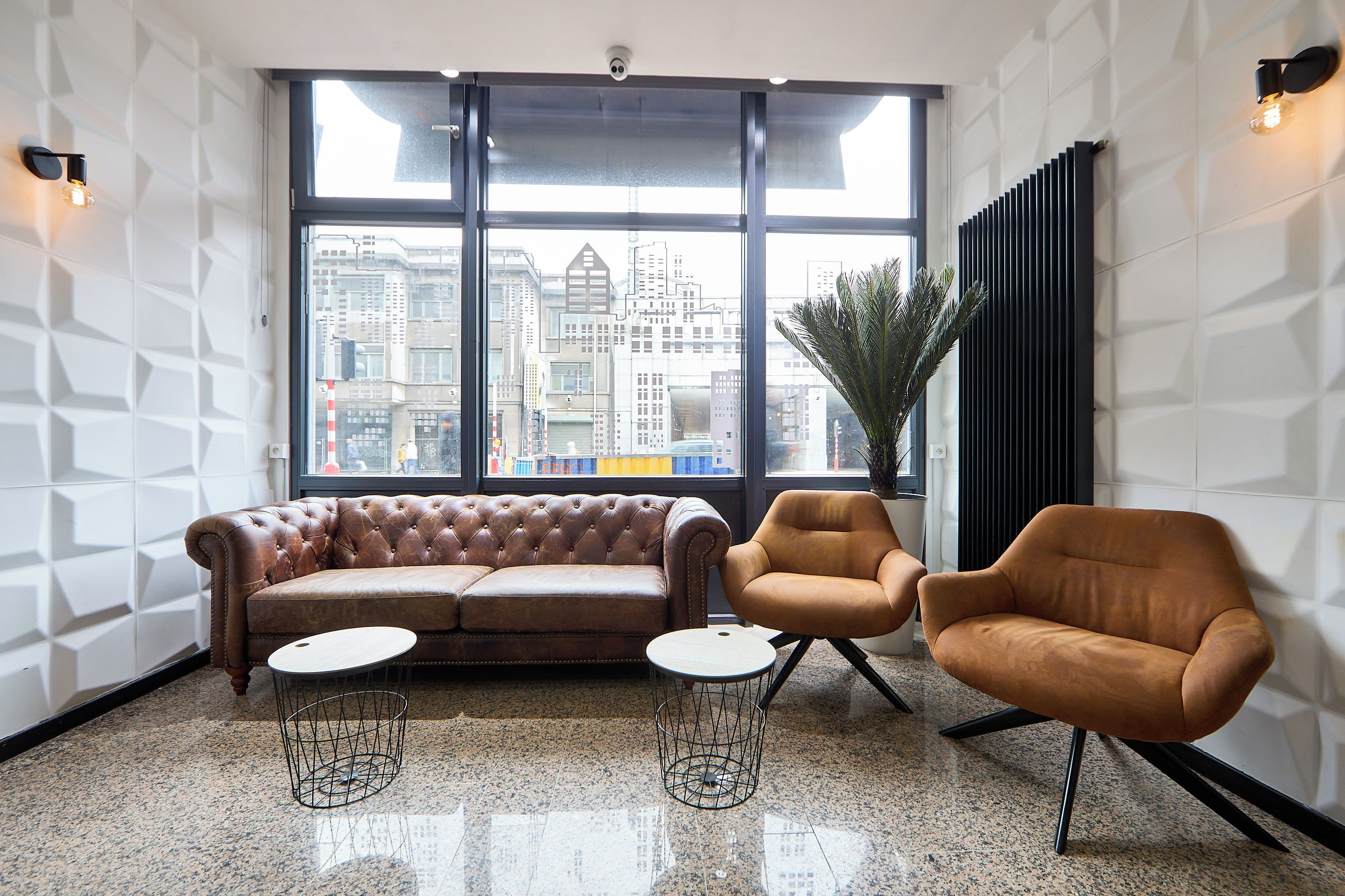 Un salón moderno con un sofá de cuero, dos sillones y una mesita. Grandes ventanales ofrecen vistas a la ciudad.