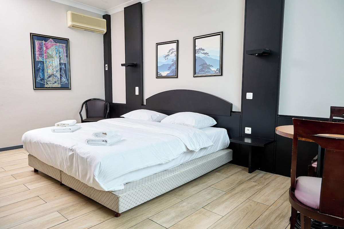 A neatly made bed with white linens and towels, a black headboard, and a small bedside table with a lamp. There are framed pictures on the wall and a wooden floor.