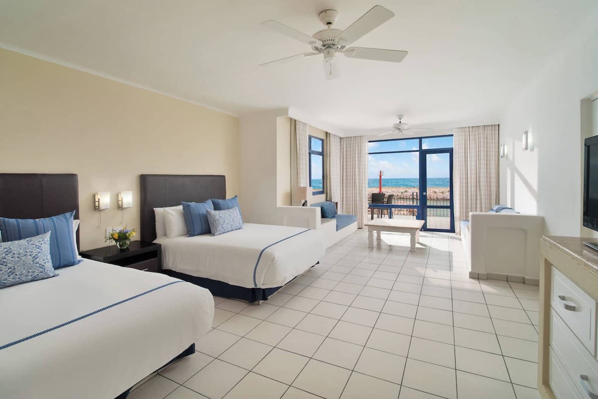 Habitación de hotel con dos camas, ventilador de techo, televisión, balcón con vista a la playa y zona de comedor.