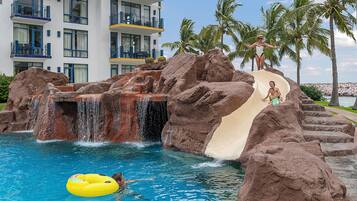 Zona de piscina con tobogán de agua, palmeras y un edificio con balcones.