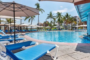 A poolside area with lounge chairs and umbrellas, surrounded by palm trees and buildings.