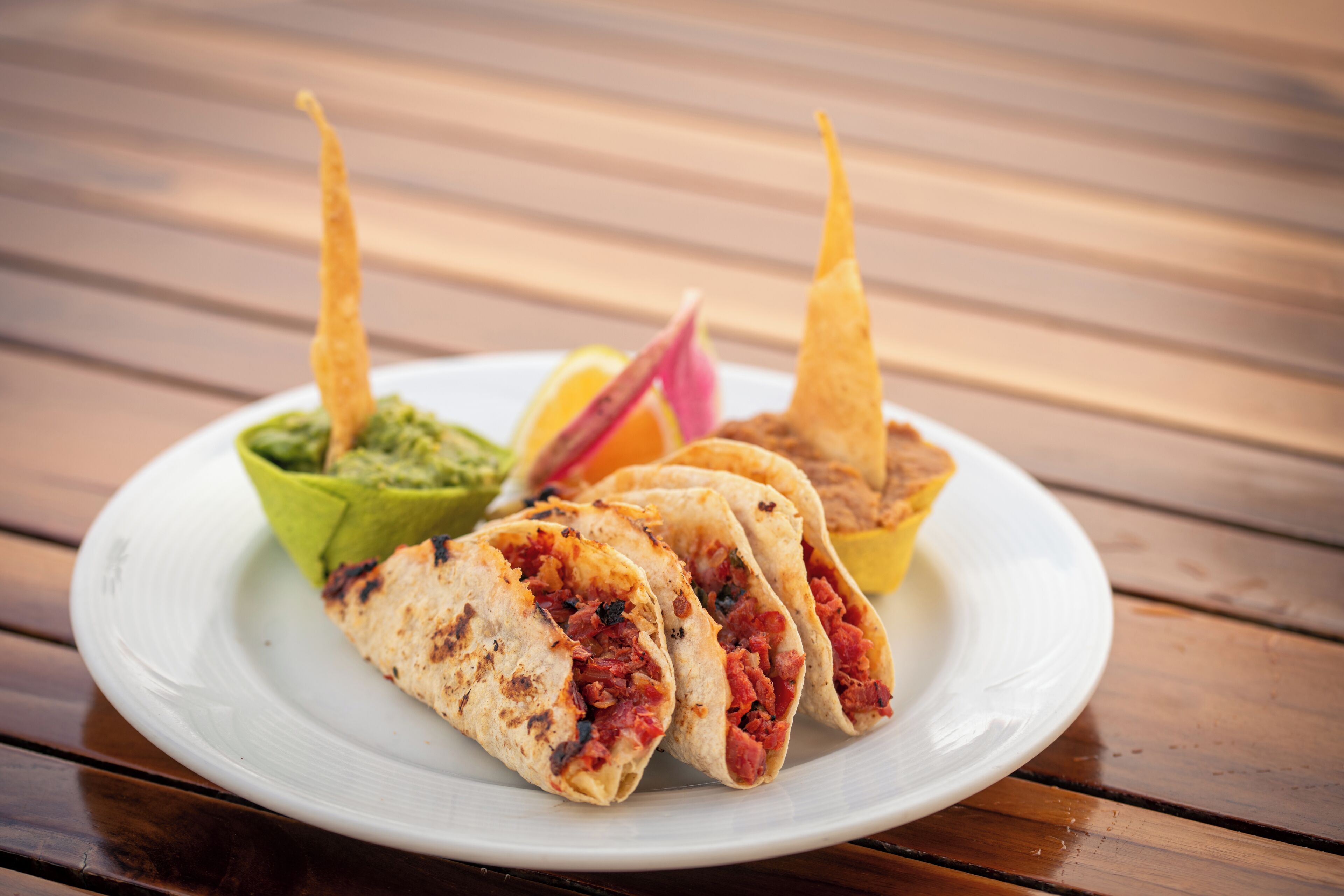 A plate with three tacos, a bowl of guacamole, and a slice of lime on a wooden table.