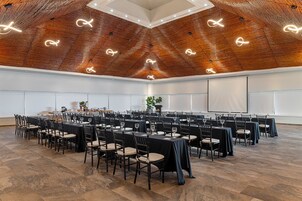 Una sala de conferencias con techo alto, mesas dispuestas y sillas, y una pantalla de proyector.