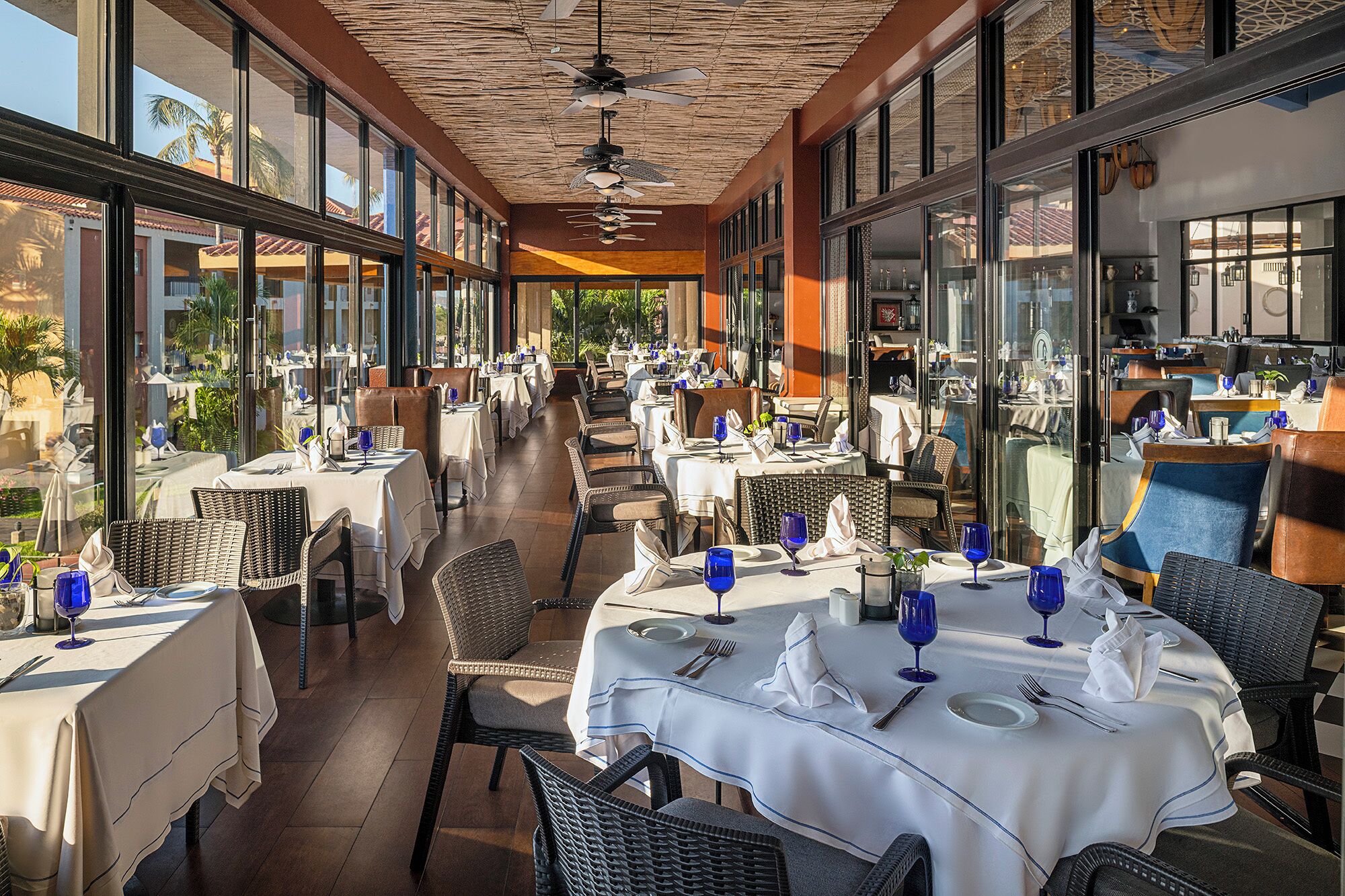 Un área de comedor al aire libre con mesas, sillas, ventanas amplias y un ventilador de techo.