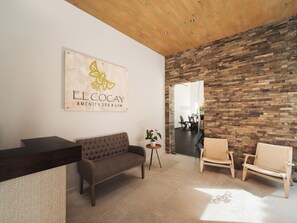 A reception area with a wooden reception desk, a brown sofa, two wicker chairs, a stone wall, and a sign reading 'EL COCAY AMENITY SPA & GYM'.