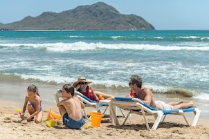 Escena de playa con gente descansando en tumbonas, un niño jugando con juguetes de arena y una montaña al fondo.