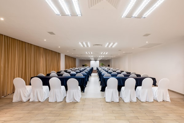 A conference room with rows of tables and chairs, a wooden panel wall, and ceiling lights.