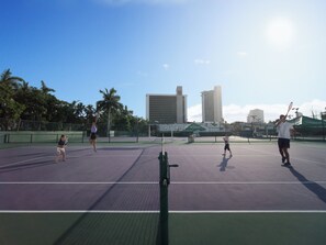 Un campo da tennis all'aperto con rete, circondato da una recinzione, e edifici sullo sfondo.