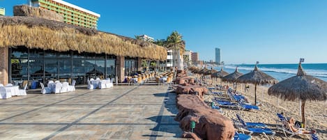A beachside hotel with outdoor seating, thatched umbrellas, and a view of the ocean.