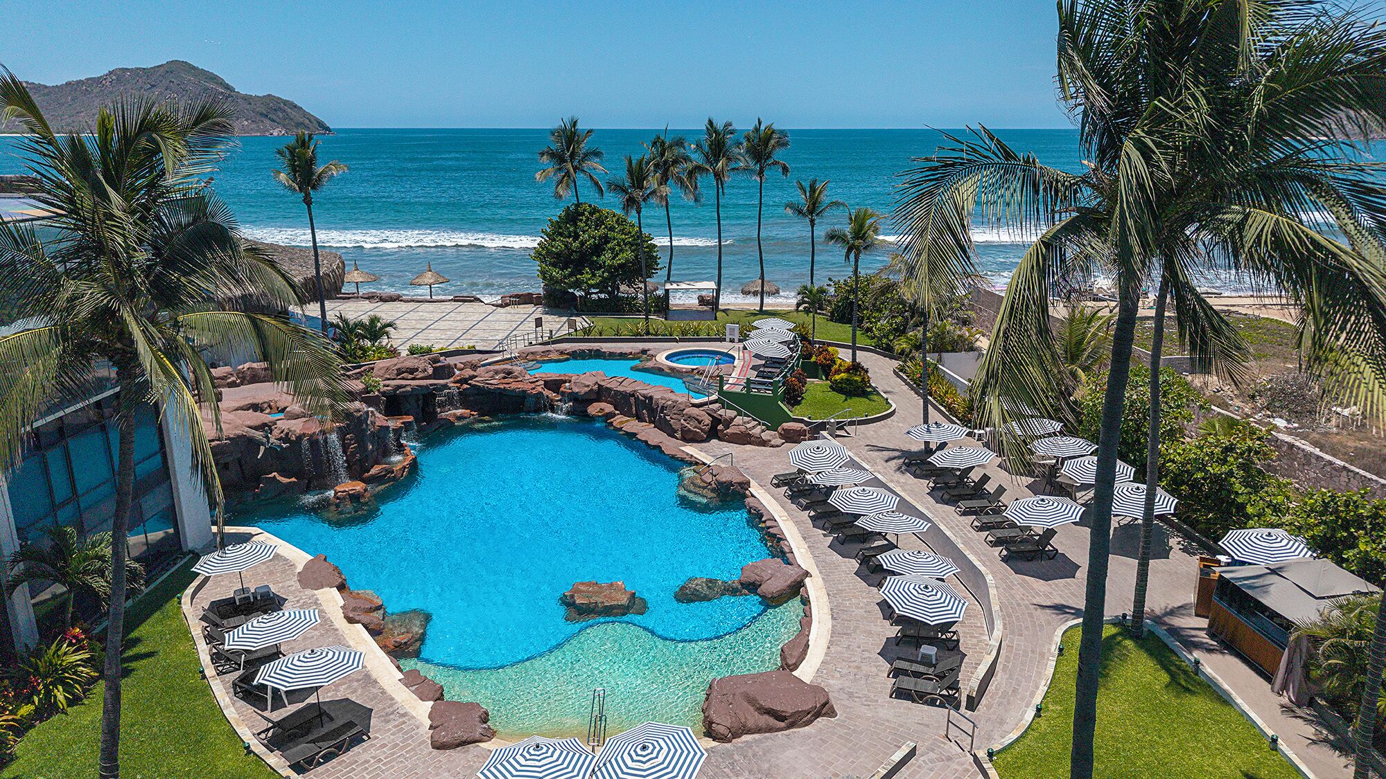 Zona de piscina en un resort con varias piscinas, sillones de descanso y sombrillas, con vista a una playa y al océano.