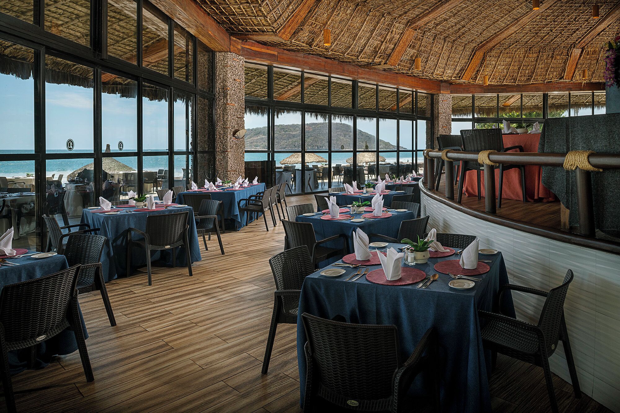 A restaurant with multiple tables set for dining, large windows offering a view of the sea, and a thatched roof ceiling.
