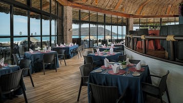 A restaurant with multiple tables set for dining, large windows offering a view of the sea, and a thatched roof ceiling.