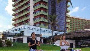 Two individuals meditating on a grassy lawn with a modern building and a thatched-roof structure in the background. - El Cid El Moro Beach Hotel (Mazatlán)